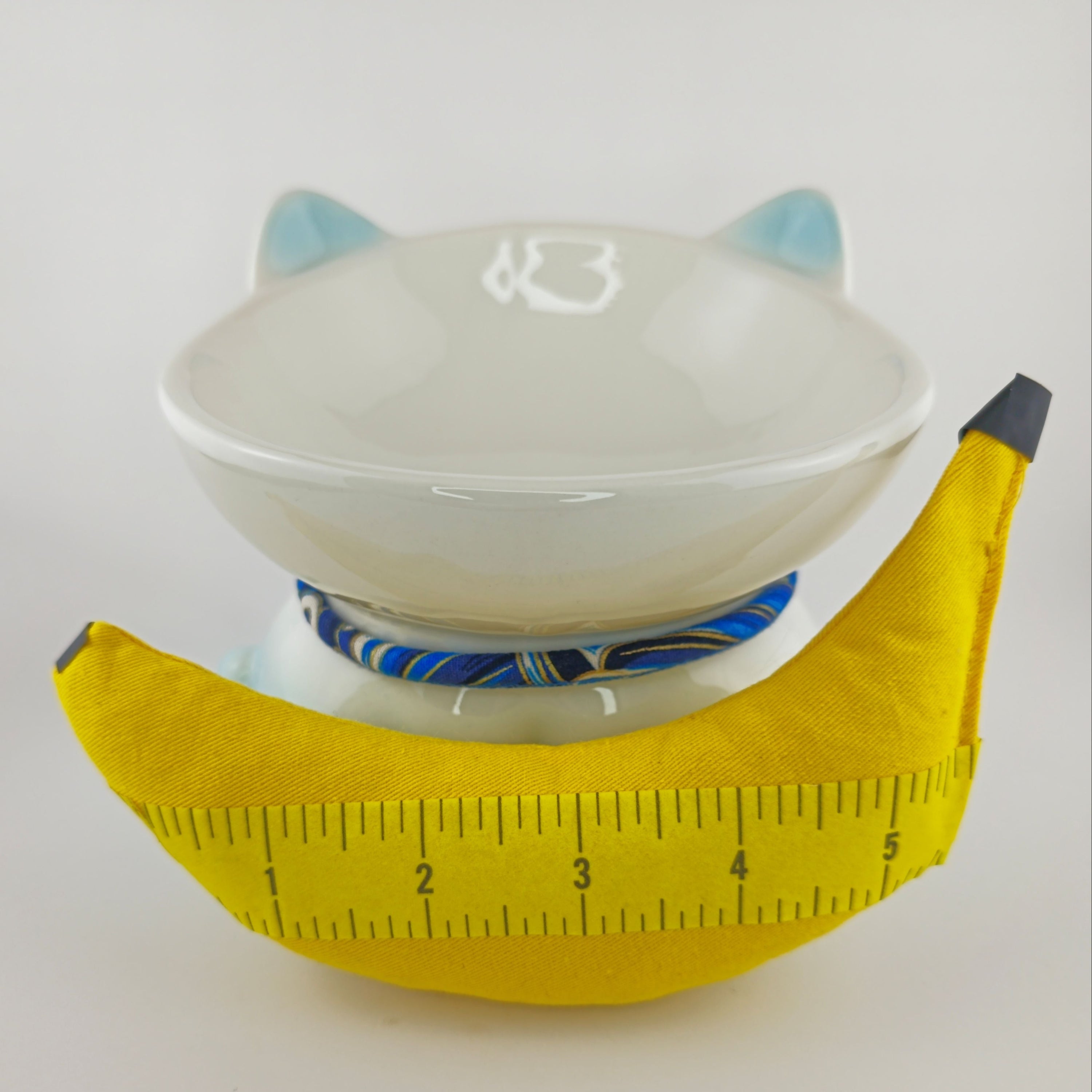 White bowl with blue interior and cat ears on a yellow banana, measured with a tape measure.
