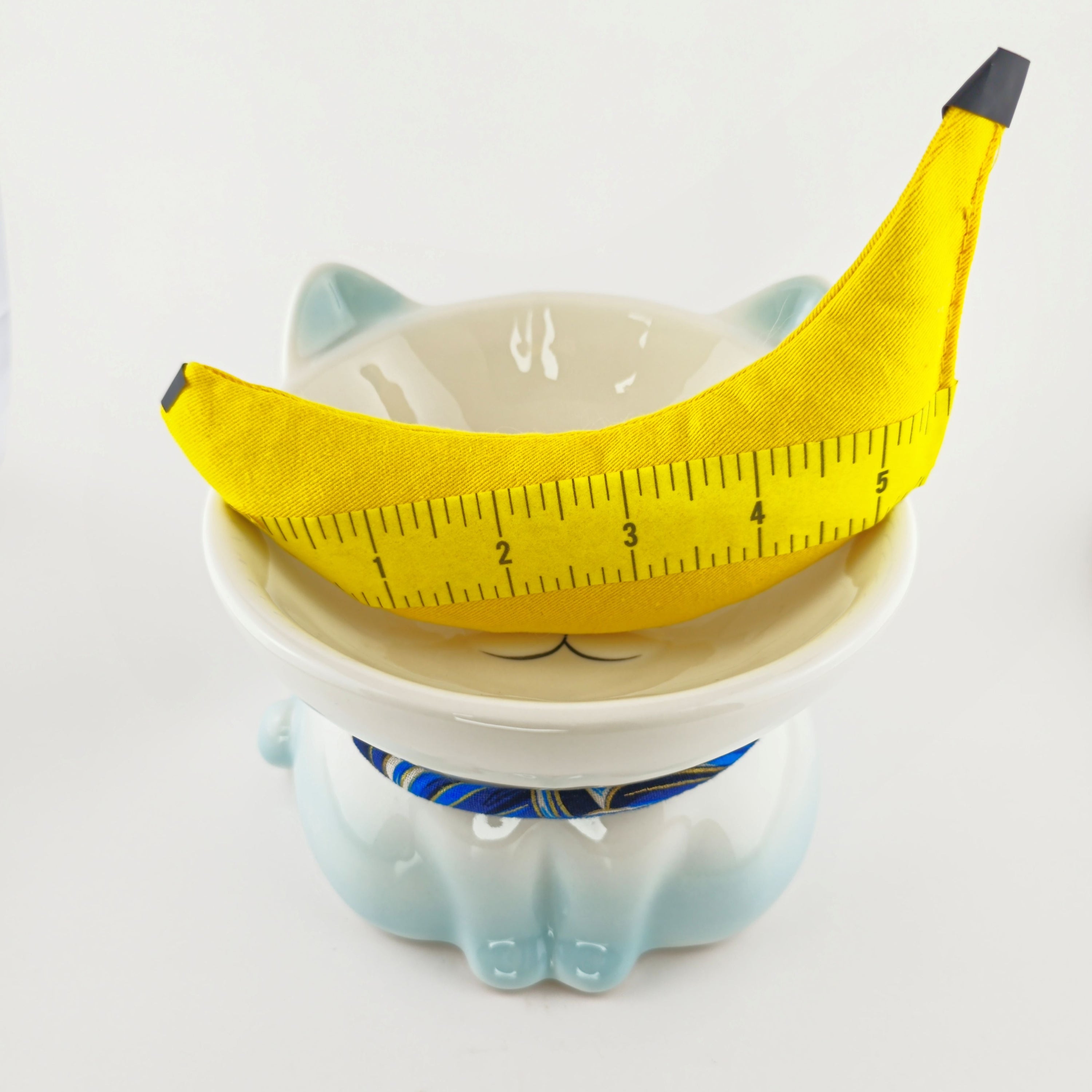 Ceramic cat-shaped bowl with a banana and measuring tape on top against a white background